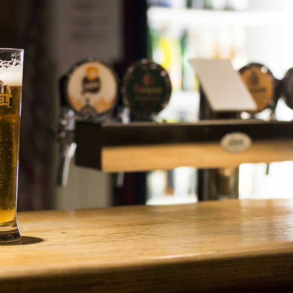 Tall glass of golden Kasteelbier rests on a wooden bar counter, with beer taps blurred in the background.