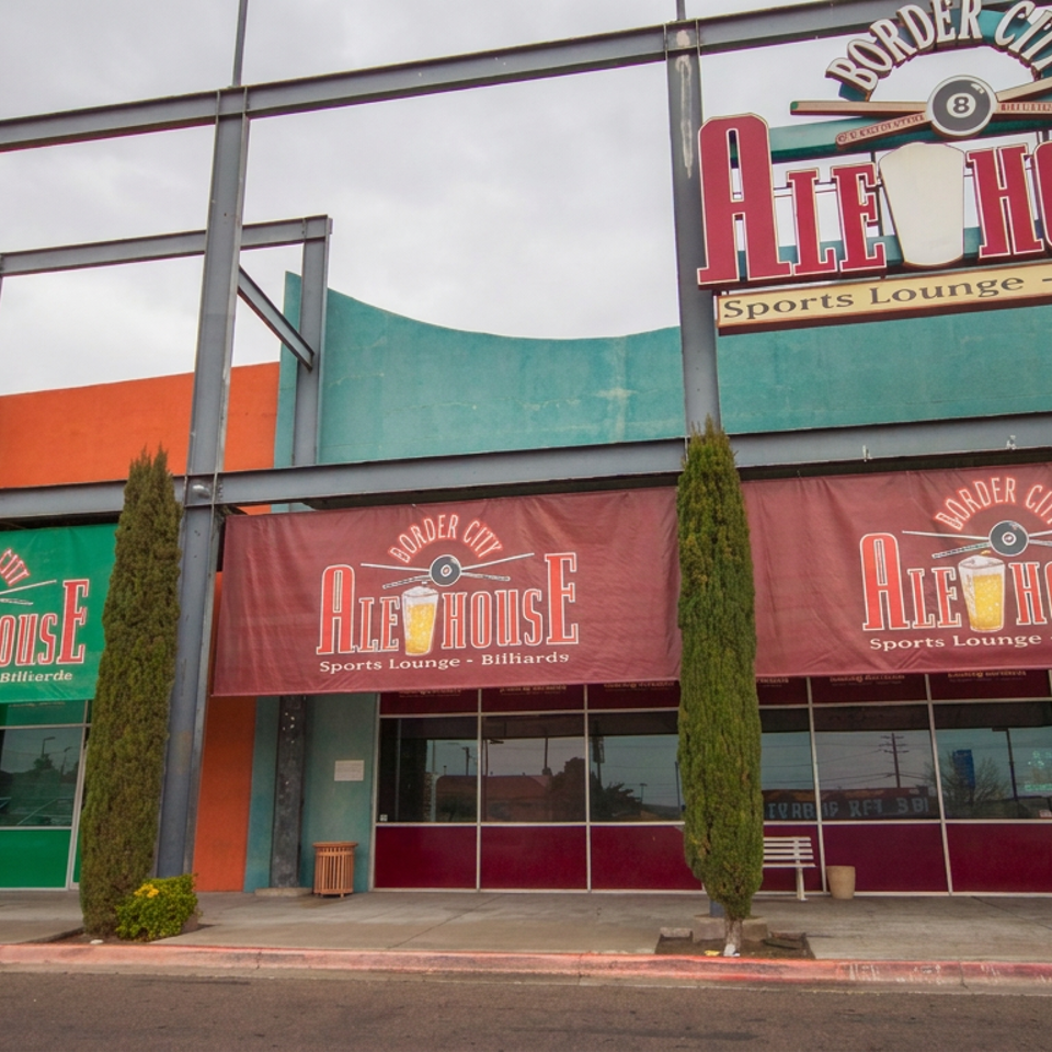 Exterior of Border City Ale House sports lounge and billiards venue with colorful awnings, signage, storefront windows, and tall shrubs.